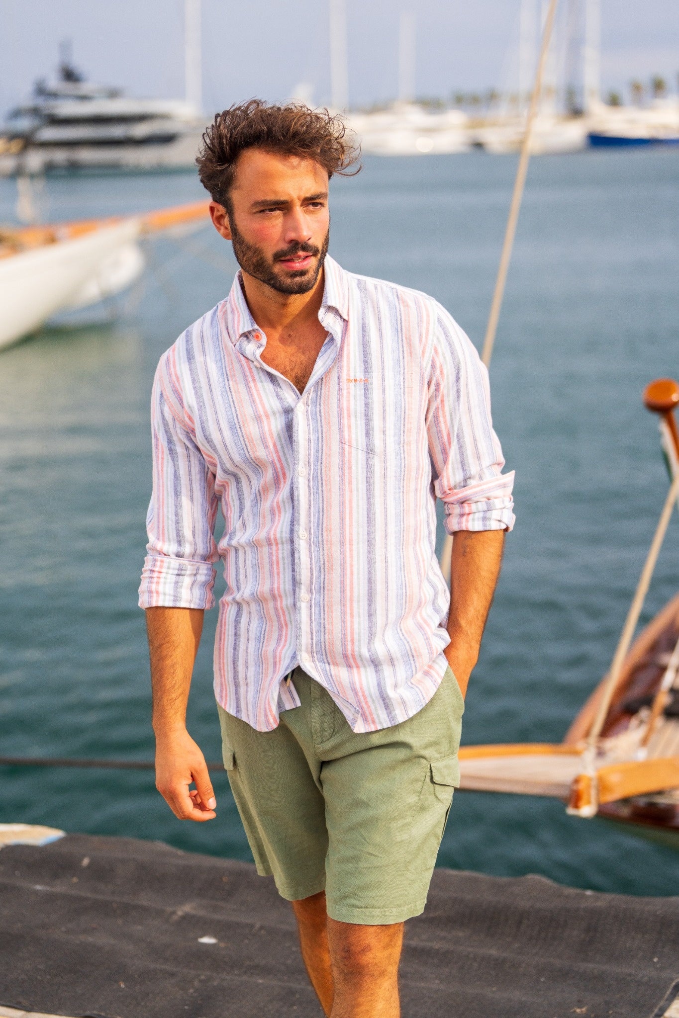 Man in a striped shirt and green shorts walking near a harbor with yachts in the background.