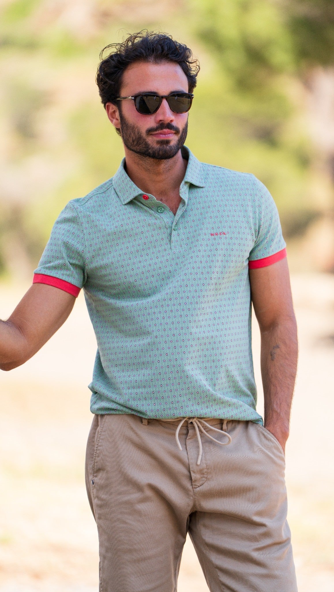 Man in a colorful plaid shirt and shorts smiling while walking outdoors near a boat and rocky backdrop.
