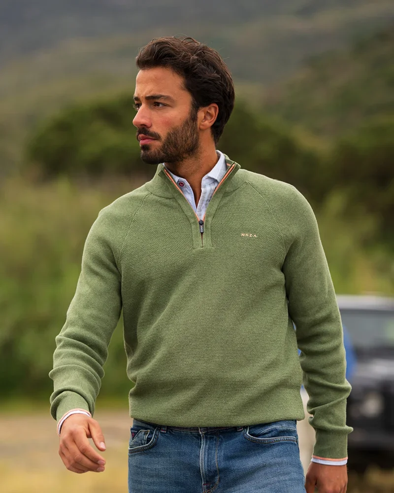 Man in a green sweater with a quarter-zip stands outside against a blurred natural background, looking to the side.