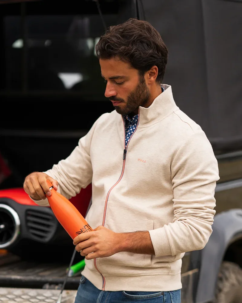 Man in a beige zip-up jacket holding an orange water bottle outdoors near a vehicle.